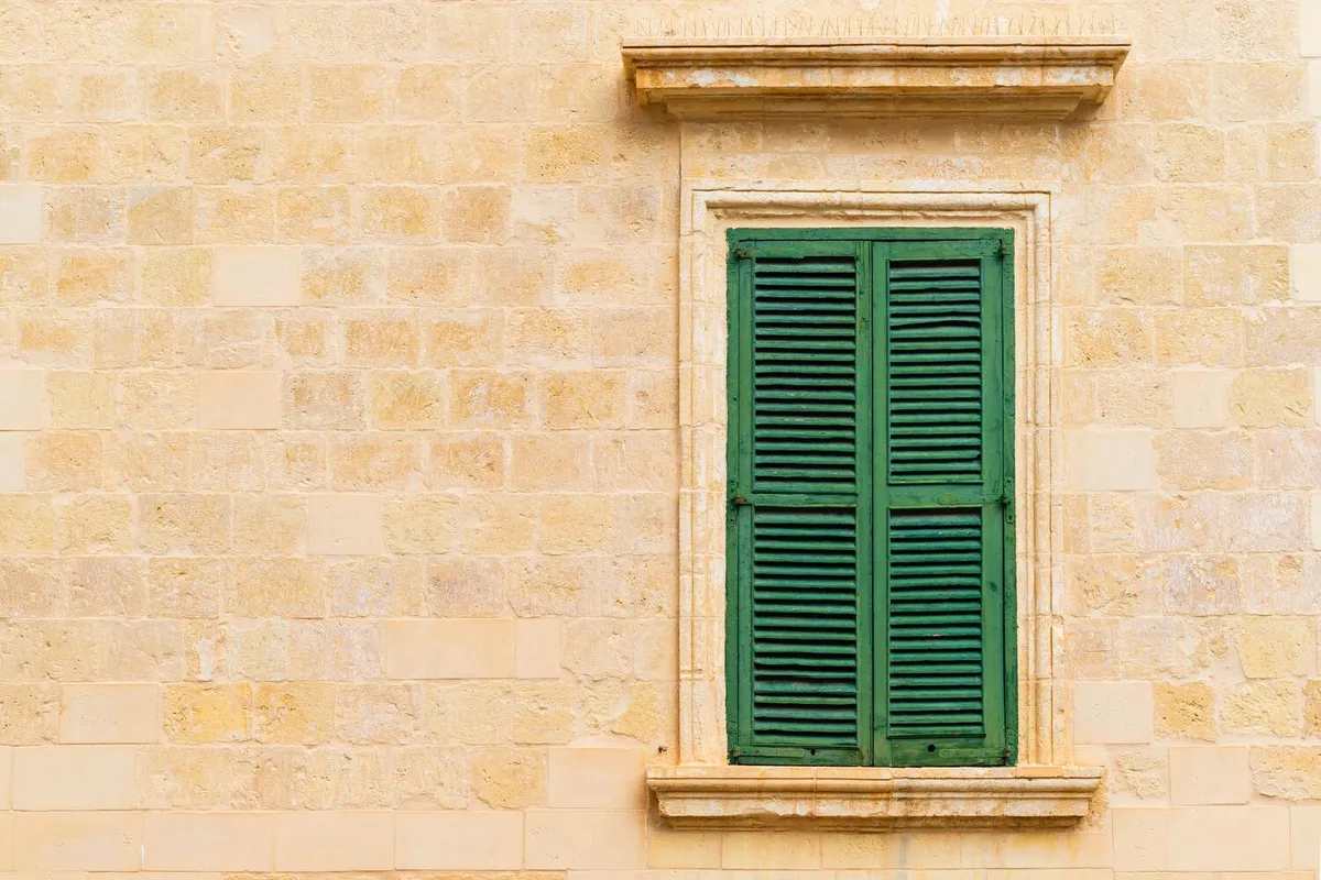 Fenêtre à persiennes en bois peintes en vert, à deux vantaux fermés, dans un mur en pierre de taille blonde