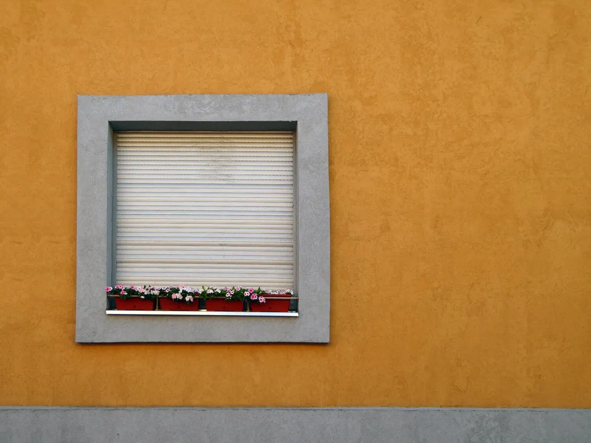 Volet roulant blanc fermé dans une fenêtre à encadrement gris, sur une façade enduite ocre avec jardinière fleurie